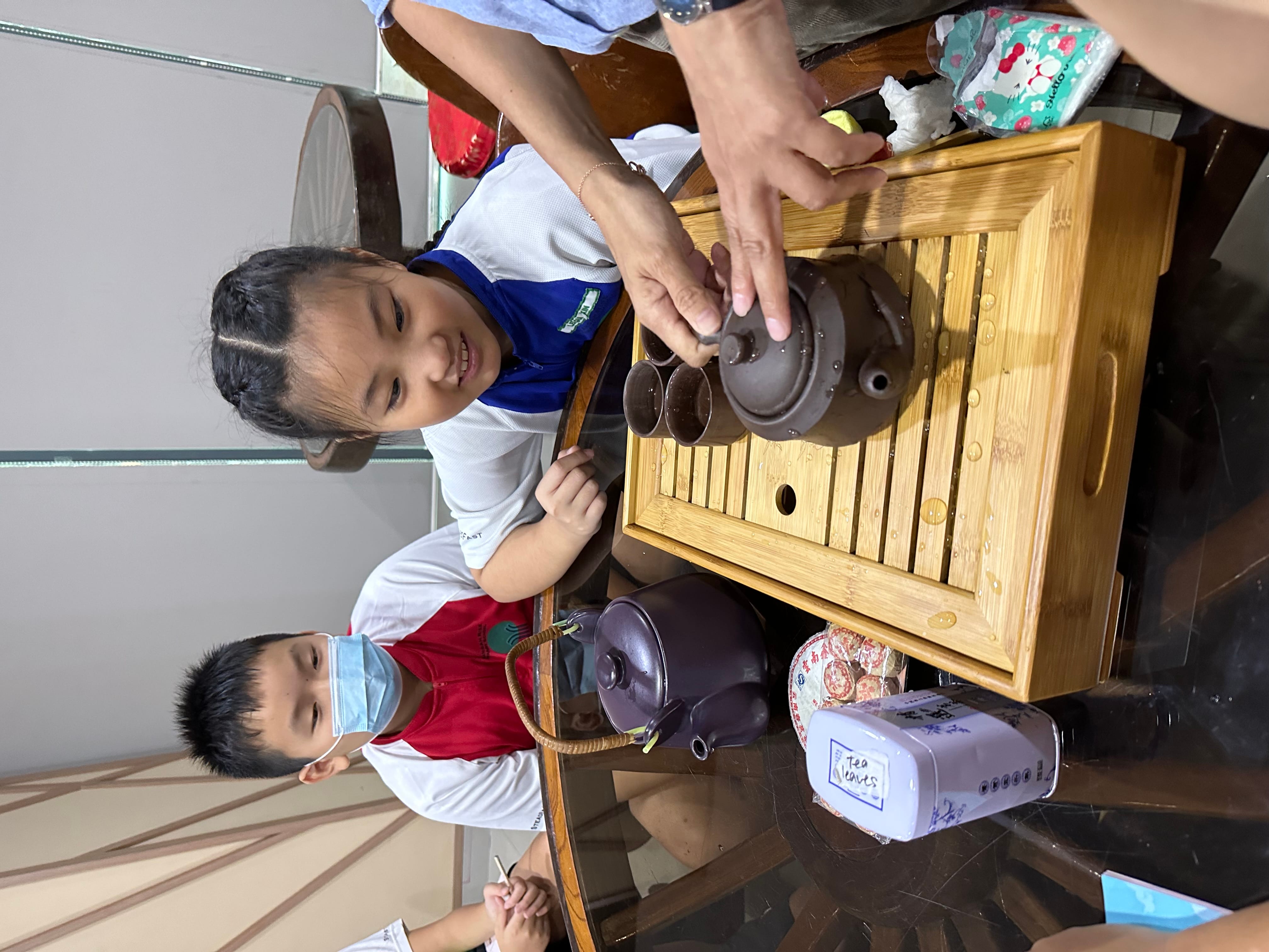 Two children sit at a table with a bamboo tea tray holding clay teapots and cups during a tea ceremony.